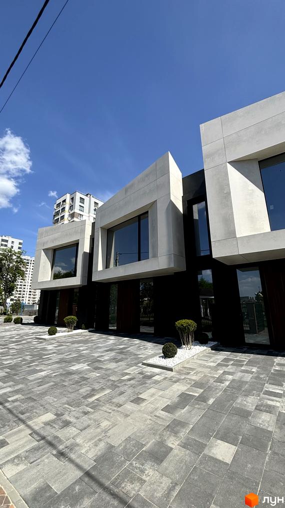 Фасад будівлі з великими вікнами та бетонним оздобленням. Перед будинком розташована бруківка та декоративні кущі.