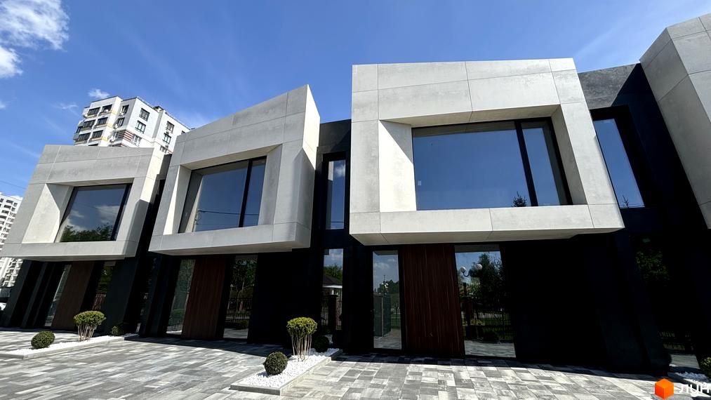 Фасад будинку з великими вікнами та світлими бетонними блоками. Перед будинком розташована бруківка та невеликі кущі.