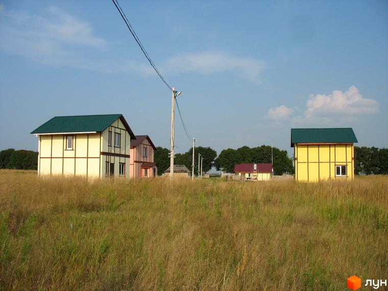 Будинки в Котеджному містечку Під Горою з жовтими та бежевими стінами, зеленими дахами та вікнами. На передньому плані суха трава, на задньому - дерева та блакитне небо.