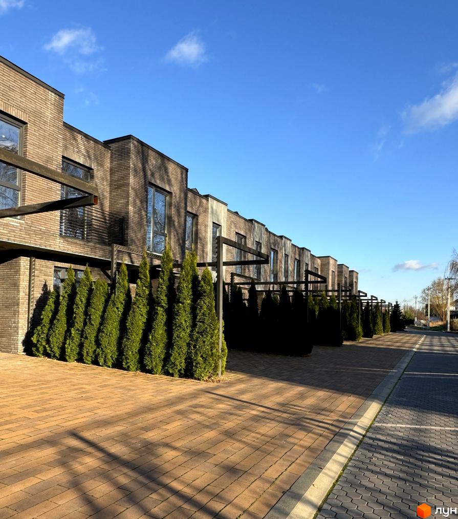Ряд двоповерхових будинків з коричневої цегли з великими вікнами. Перед будинками висаджені зелені туї, а територія викладена коричневою плиткою.