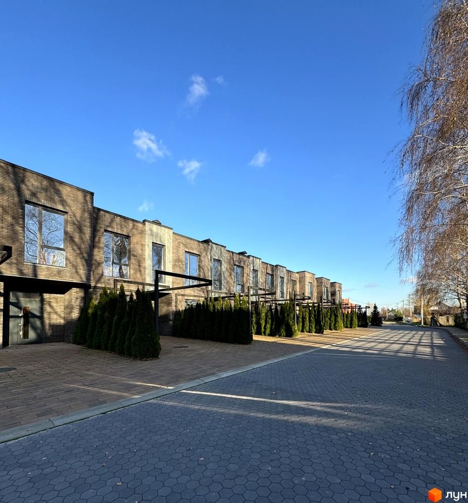 Ряд будинків з коричневої цегли з великими вікнами та чорними навісами. Перед будинками висаджені туї, а дорога викладена плиткою.