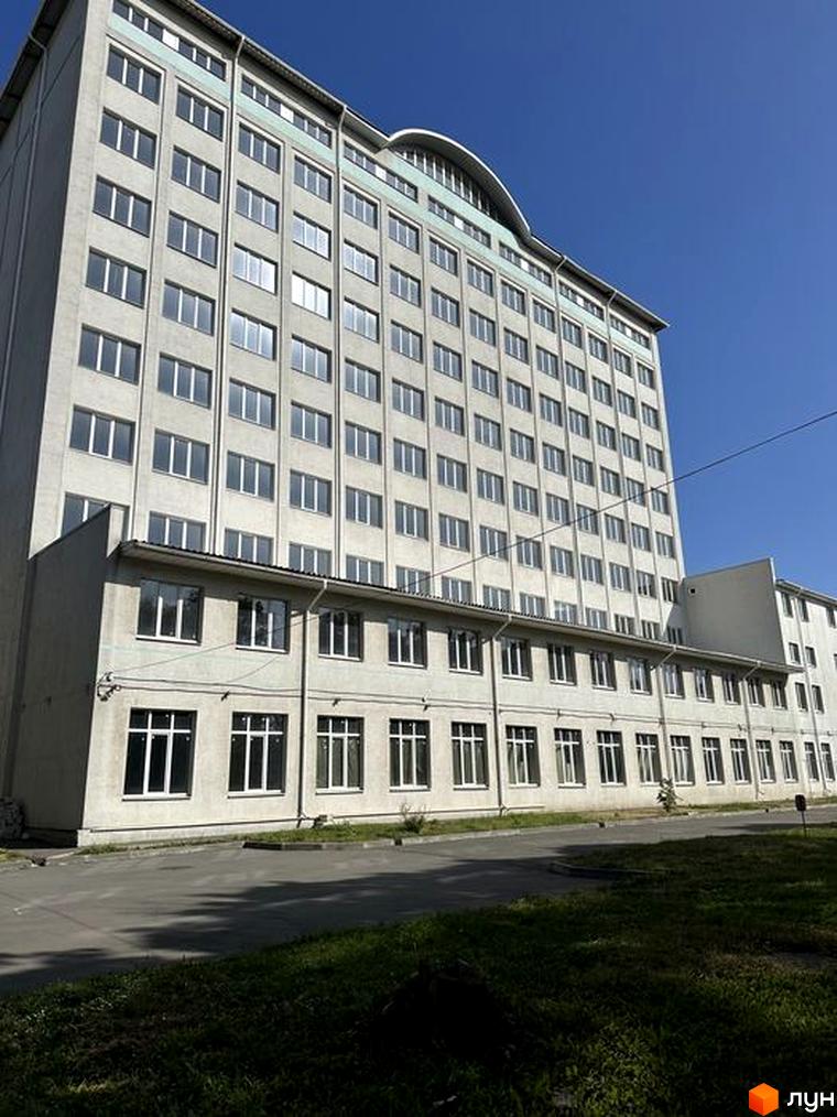 Висотна будівля з великою кількістю вікон. Перед будинком асфальтована дорога та зелена зона.