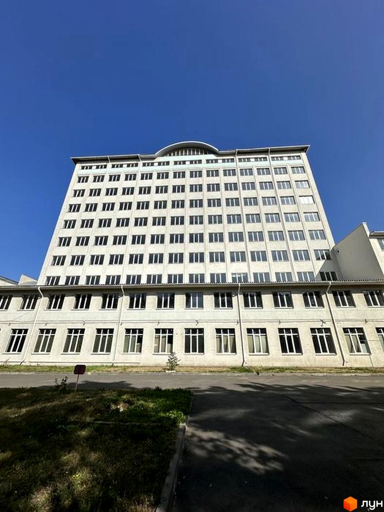 Висока будівля з великою кількістю вікон на фоні блакитного неба. Перед будівлею розташована зелена зона та асфальтована дорога.