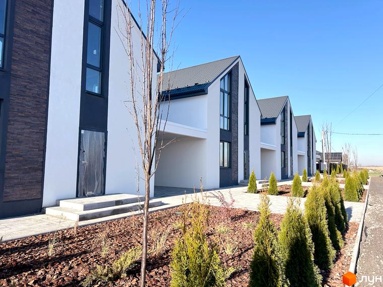 Котеджне містечко DESNA HOMES з білими будинками з темними вставками та великими вікнами, перед будинками висаджені туї та декоративні рослини.