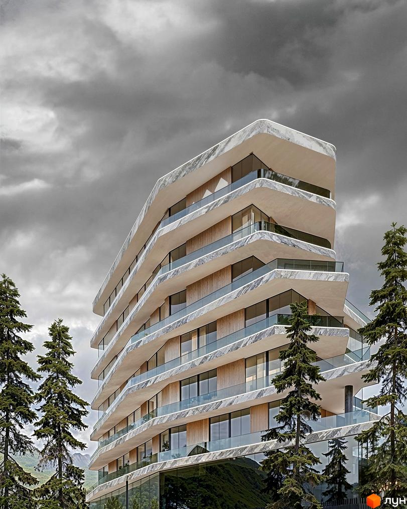 Багаторівнева будівля ZAGRAVA Villas & Residence з хвилястими фасадами, облицьованими світлим каменем, та скляними балконами, оточена високими хвойними деревами на тлі гір.