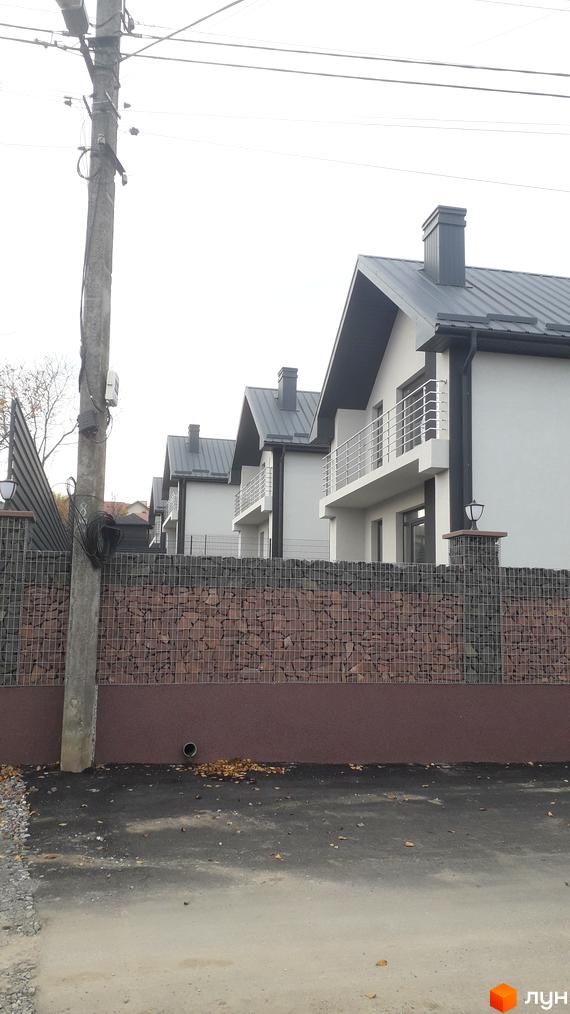 Ряд котеджів з білими фасадами, сірими дахами та балконами. Перед будинками паркан з габіонів та коричневою основою.