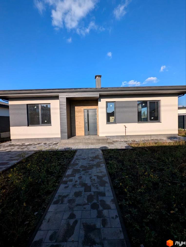 Одноповерховий будинок світло-бежевого кольору з темно-сірими вікнами та елементами фасаду. До будинку веде вимощена доріжка, поруч зелена ділянка.