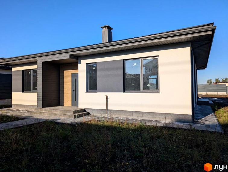 Одноповерховий будинок з темно-сірим дахом та світло-бежевими стінами з темними акцентами. До входу веде вимощена доріжка, навколо ділянка з травою.