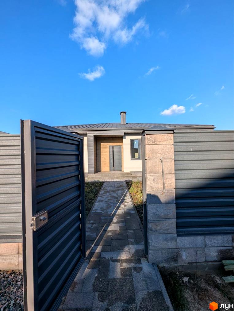 Одноповерховий будинок з сірим дахом та світлими стінами, дерев'яним оздобленням біля входу. До будинку веде вимощена доріжка через відкриту металеву браму та огорожу.