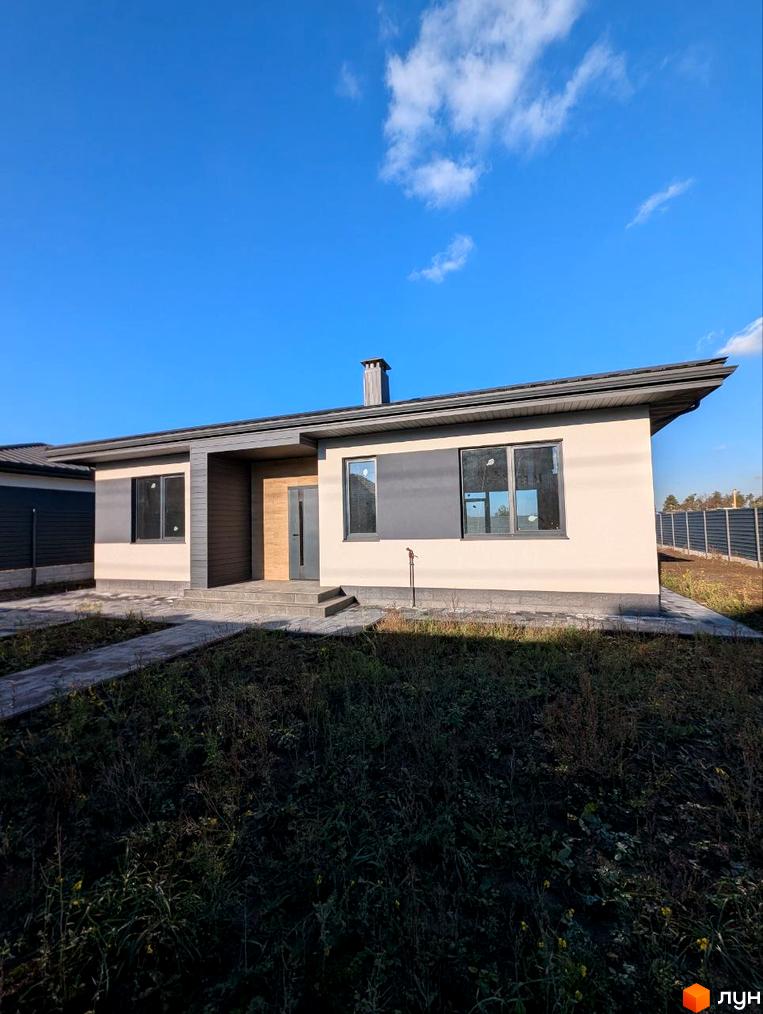 Одноповерховий будинок світлого кольору з темними віконними рамами та дахом. Навколо будинку вимощені доріжки та ділянка з травою.