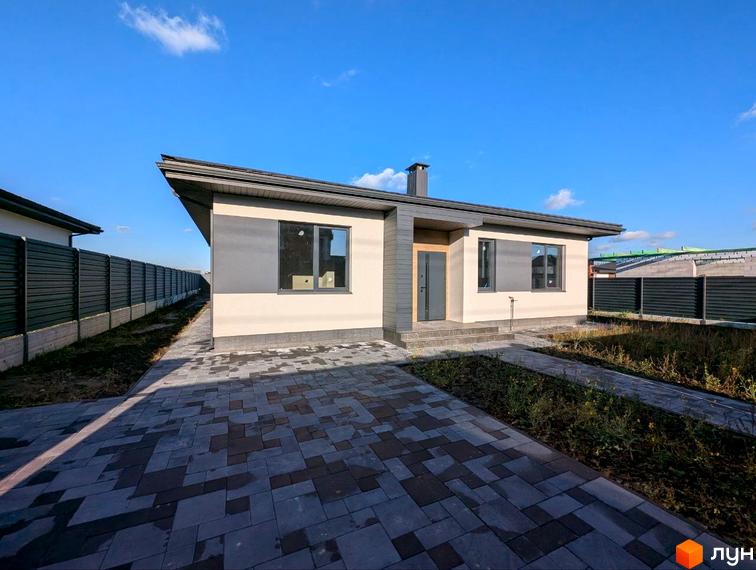 Одноповерховий будинок світлого кольору з темними акцентами та похилим дахом. Перед будинком вимощена доріжка з тротуарної плитки та зелена ділянка біля огорожі.