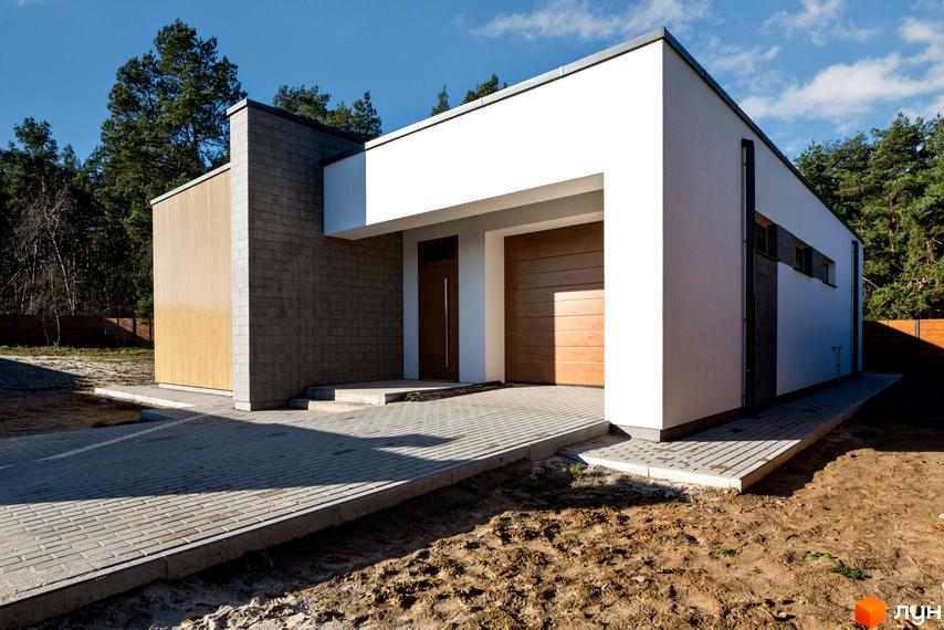 Одноповерховий будинок Котеджного містечка LOTUS з білими стінами, цегляними та дерев'яними елементами. Перед будинком вимощені доріжки, навколо ділянка з ґрунтом та сосни.