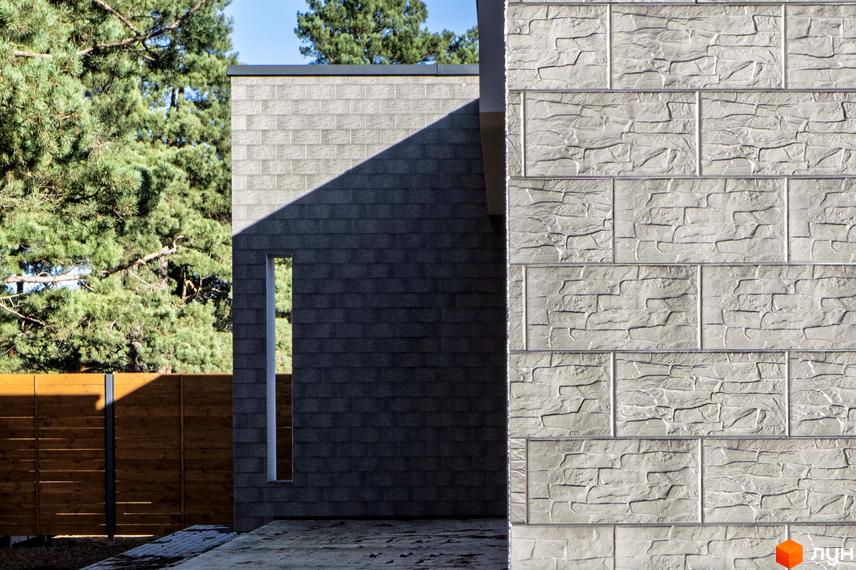 Котеджне містечко LOTUS: фасад будинку з рельєфними світлими панелями та темною цегляною кладкою з вузьким вікном. Навколо високі сосни та дерев'яний паркан.