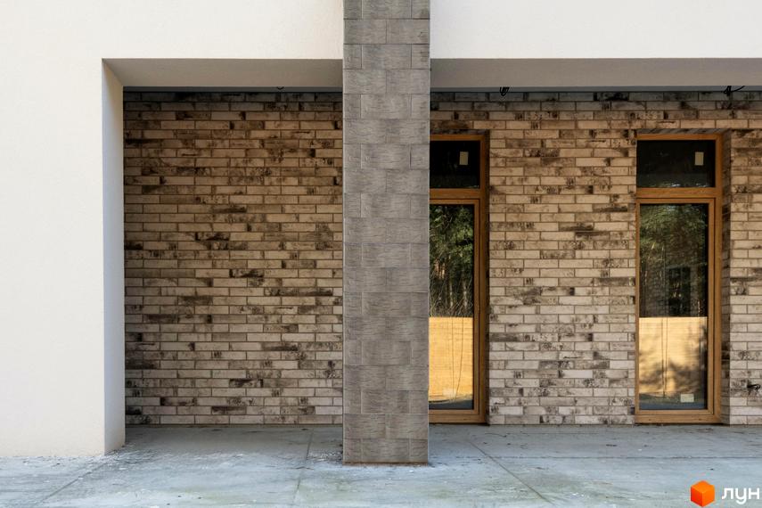 Фасад будинку Котеджного містечка LOTUS з цегли світлих відтінків, сірою колоною та великими вікнами. Перед будинком бетонна тераса.