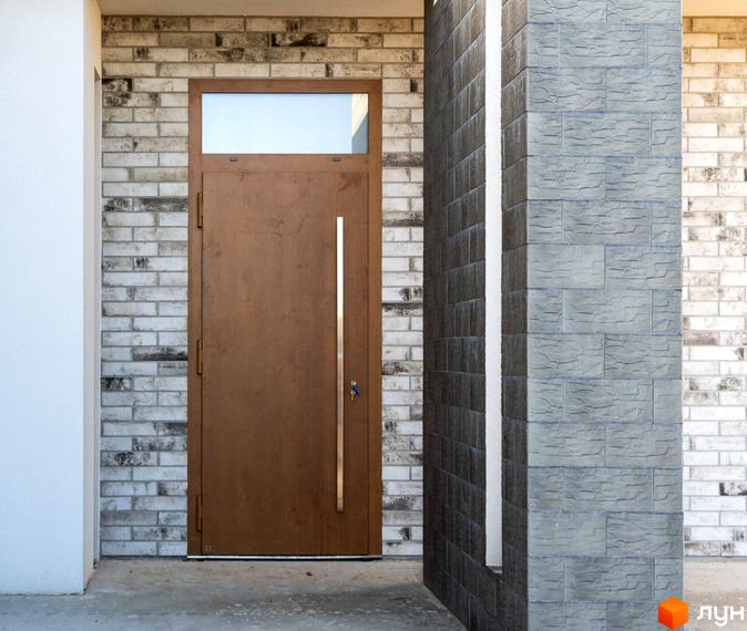 Вхід до будинку Котеджного містечка LOTUS з коричневими дверима, вікном та фасадом з цегли світлих відтінків, поруч колона з сірих плиток.
