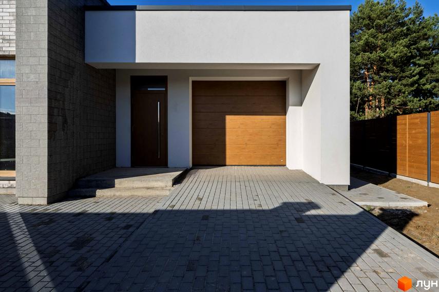 Котеджне містечко LOTUS: білий фасад будинку з темно-коричневими дверима та дерев'яними гаражними воротами, вимощена бруківкою територія.