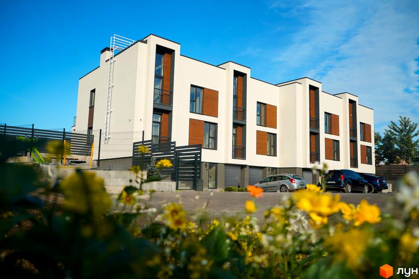 Таунхауси Motuz Hill з світлими фасадами, дерев'яними вставками та балконами. На передньому плані квіти, поруч припарковані автомобілі та дитячий майданчик.