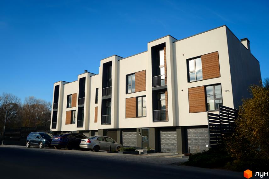 Комплекс таунхаусів Motuz Hill з бежевими фасадами, темними віконними рамами та балконами. Перед будинками розташована парковка з автомобілями та зелені насадження.