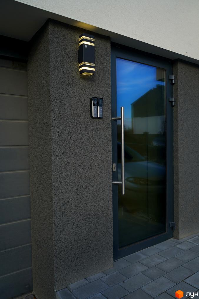 Вхід до таунхаусу Motuz Hill з темним фасадом, скляними дверима, вуличним освітленням та домофонами. Прибудинкова територія вимощена бруківкою.