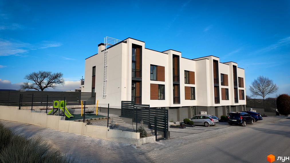 Таунхауси Motuz Hill: білі фасади з темними вікнами та дерев'яними вставками, поруч дитячий майданчик і парковка.