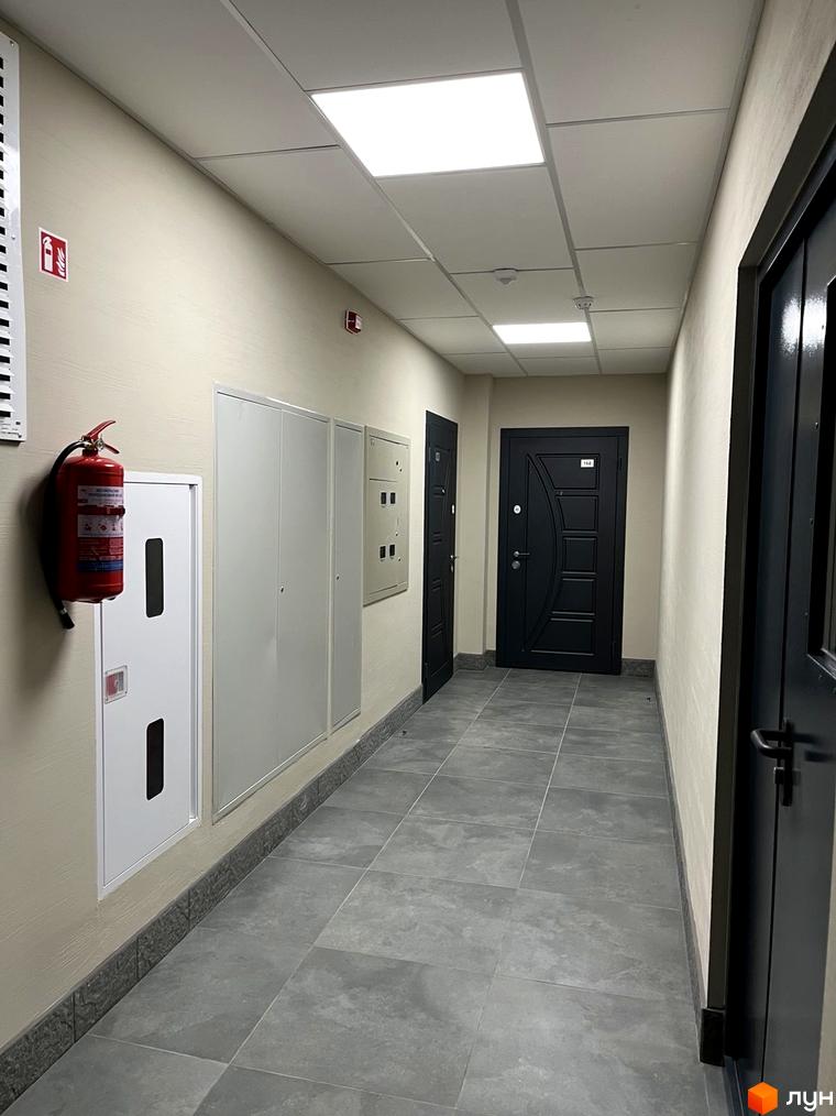 Коридор Житлового комплексу GRANIT з темними вхідними дверима, світлими стінами, сірою плитковою підлогою та освітленням.