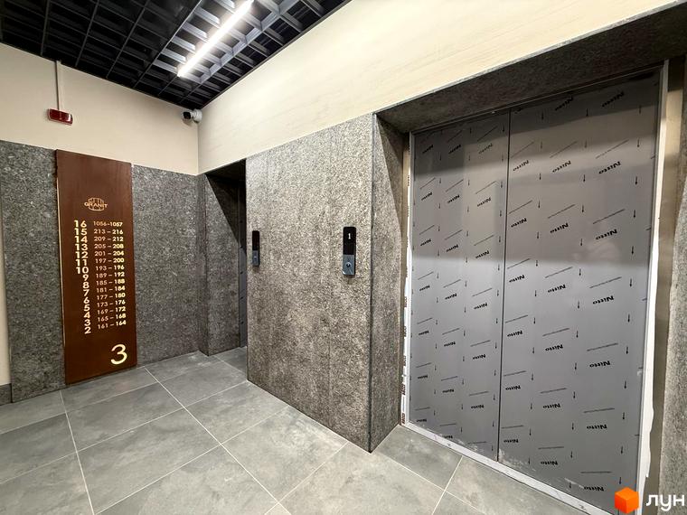 Холл ліфта Житлового комплексу GRANIT на третьому поверсі, оздоблений кам'яною плиткою, з інформаційною дошкою та дверима ліфта в захисній плівці.