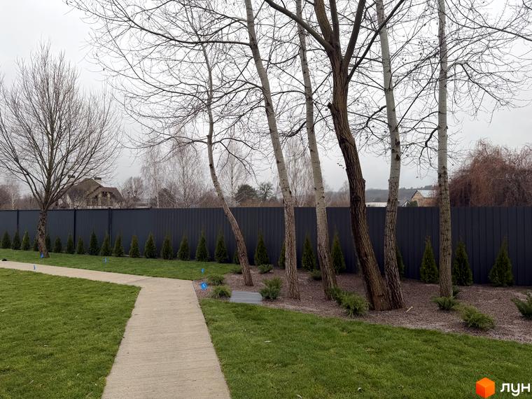 Таунхауси 11Dreams: будинок з багатоскатним дахом за темним парканом. Перед ним доріжка, газон та ряди хвойних насаджень.