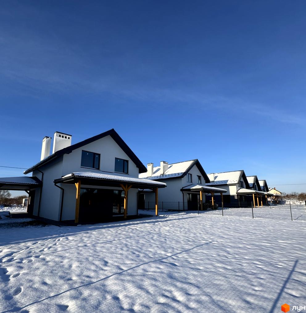 Зимовий вигляд Котеджного містечка Буде Дім. Годовиця SPA: світлі будинки з темними дахами та дерев'яними елементами, що стоять на засніженій ділянці під чистим синім небом.
