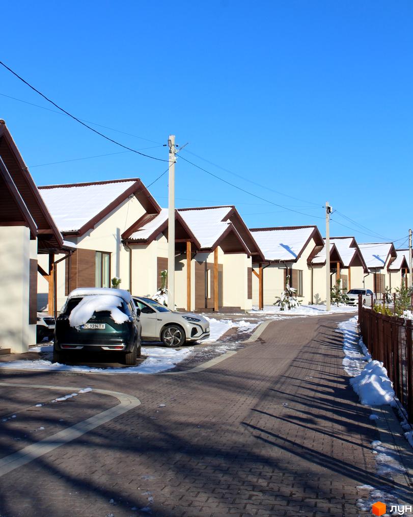 Ряд одноповерхових будинків з кремовими стінами та коричневими дахами, вкритими снігом, у Котеджному містечку Буде Дім. Деревач. Бруківка та припарковані авто на засніженій території.