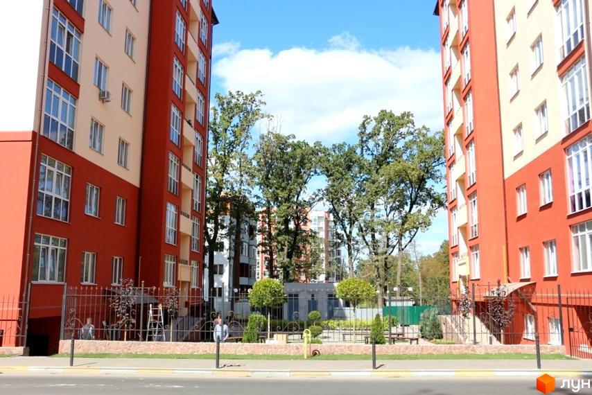 Високі будинки з червоними фасадами та світлими вставками. На прибудинковій території є зелені насадження та ковані огорожі.