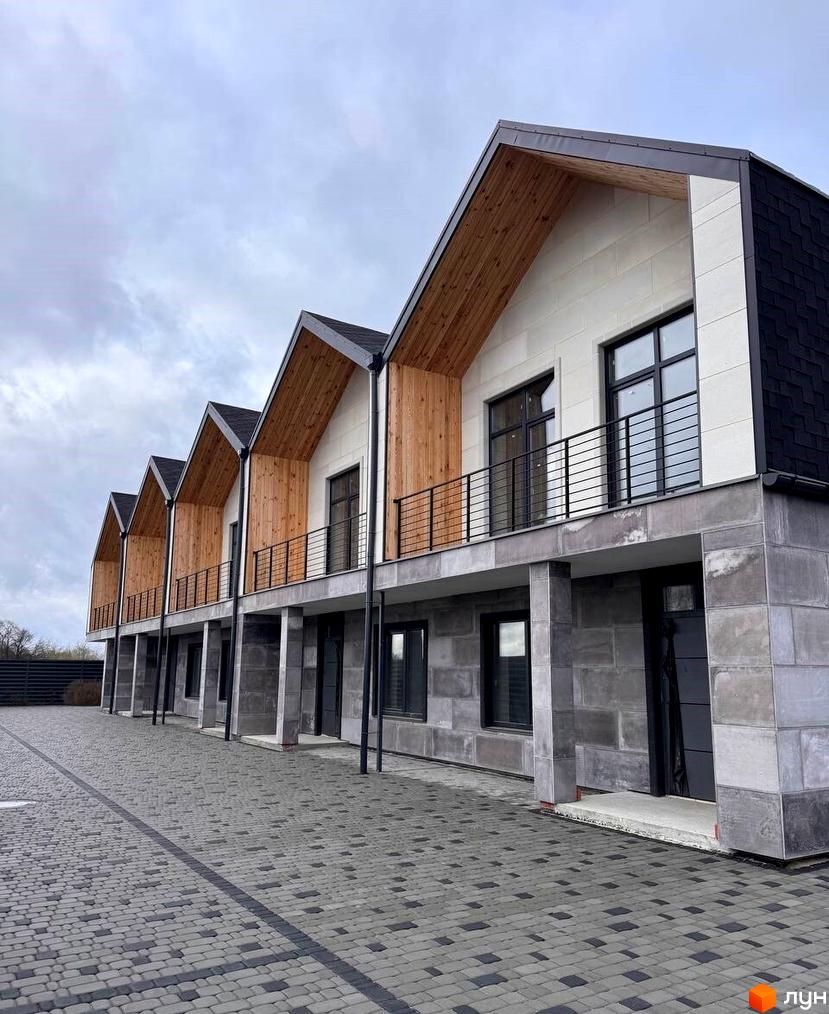 Ряд таунхаусів Riverium з чорними дахами, світлими фасадами, дерев'яними елементами та балконами. Прибудинкова територія вимощена сірою бруківкою.