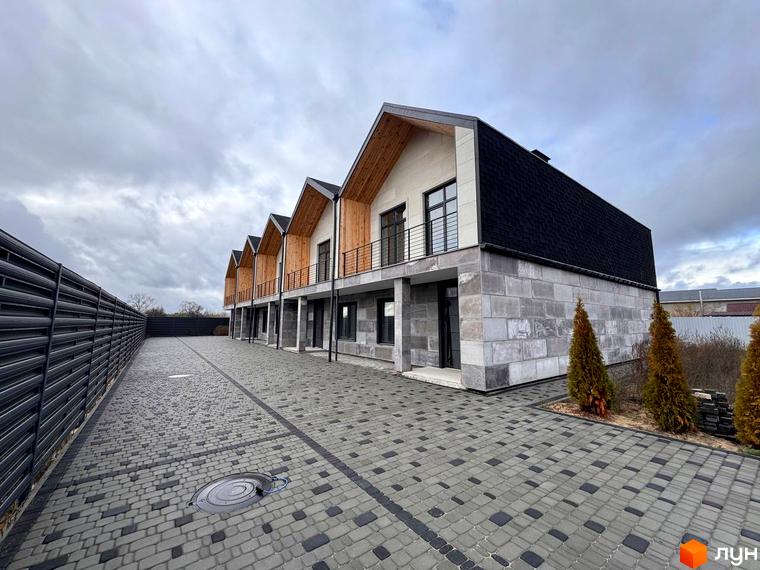 Ряд таунхаусів Riverium з темними дахами, світлими фасадами та дерев'яними елементами. Прибудинкова територія вимощена бруківкою.