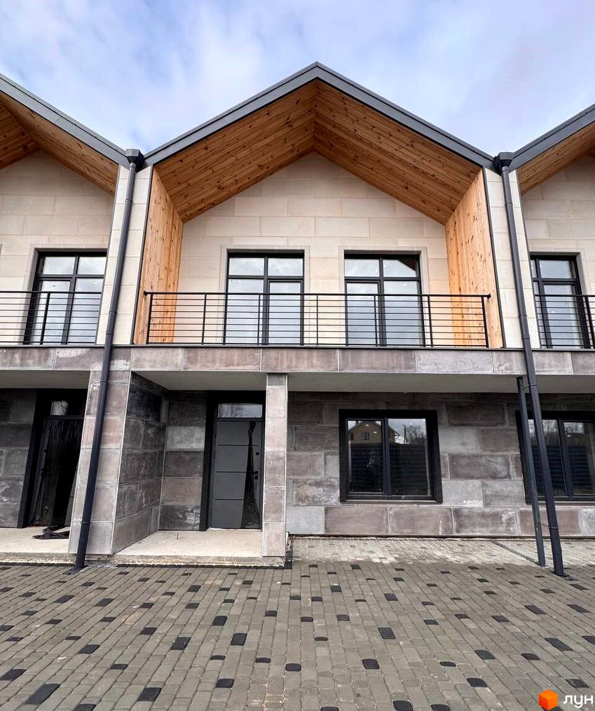 Таунхауси Riverium з фасадами з бежевих та сірих блоків, дерев'яними підшивками даху, чорними вікнами та балконами. Прибудинкова територія викладена бруківкою.