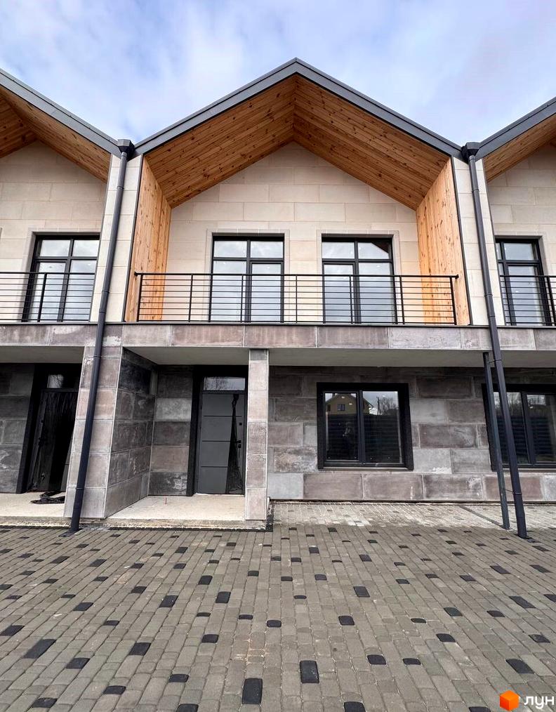 Фасад Таунхауси Riverium з бежевою та сірою обробкою, дерев'яними елементами даху та чорними вікнами. Перед будинками викладена бруківка сірого та чорного кольорів.