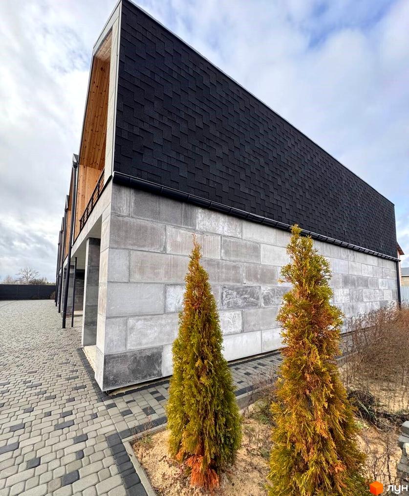 Таунхауси Riverium з темним дахом-фасадом та світло-сірими бетонними стінами. Прибудинкова територія викладена бруківкою, поруч ростуть хвойні дерева.