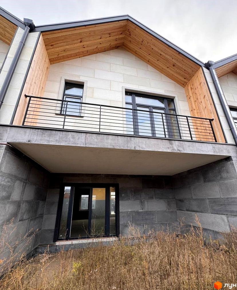 Зовнішній вигляд таунхауса Riverium зі світлим фасадом, дерев'яними вставками та балконом з металевими перилами. Прибудинкова територія з сухою травою.