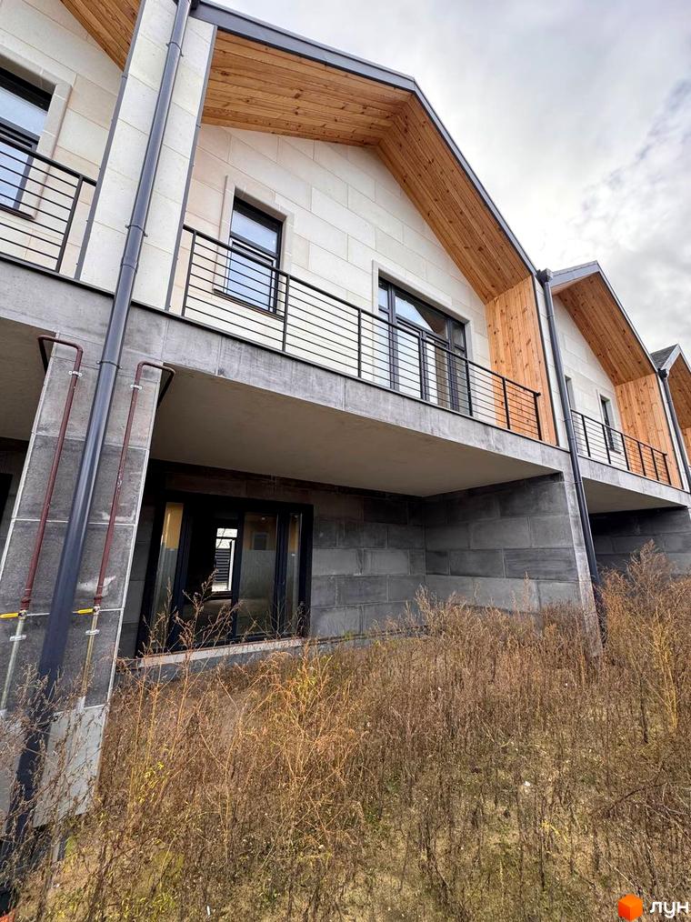 Таунхауси Riverium з світлим фасадом, дерев'яними елементами даху та чорними балконами. На передньому плані суха трава.