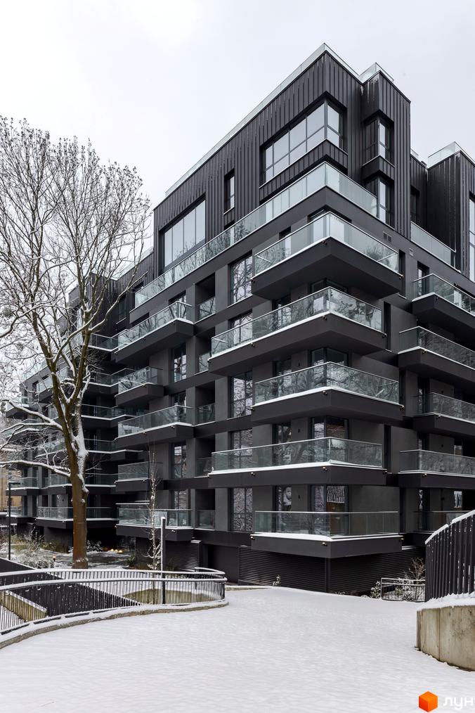 Житловий комплекс Avalon Prime: багатоповерховий будинок темного кольору зі скляними балконами та засніженою прибудинковою територією з деревами та доріжками.