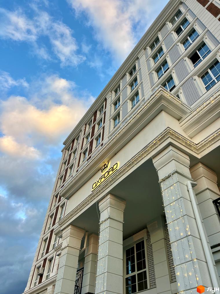 Житловий комплекс OXFORD: багатоповерховий будинок білого кольору з червоними та сірими вставками, прикрашений гірляндами на колонах.