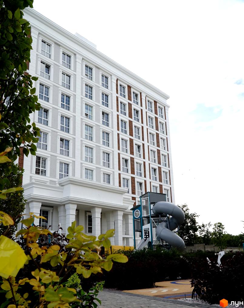 Багатоповерховий будинок Житлового комплексу OXFORD з білим фасадом та вікнами. Поруч дитячий майданчик з гіркою та зелені насадження.