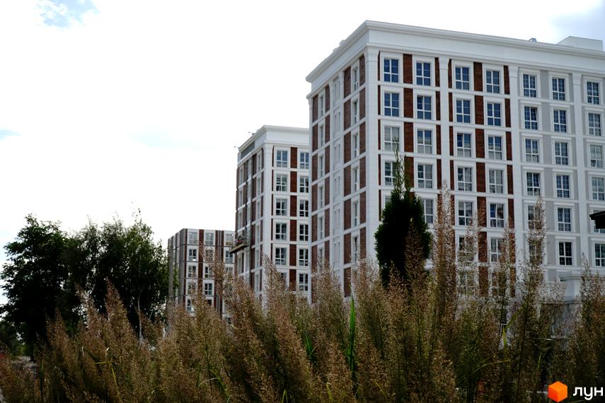 Житловий комплекс OXFORD з білими фасадами та коричневими вставками навколо вікон. Перед будинками видно високу суху траву та зелені дерева.