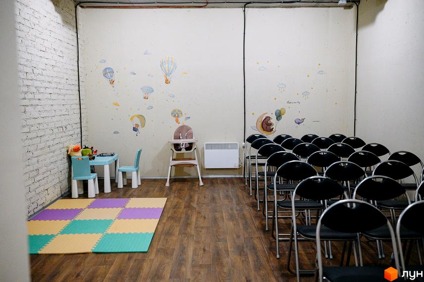 Дитяча кімната з ігровим килимком, столом та стільцями, а також зоною для проведення заходів з розкладними стільцями у Житловий комплекс OXFORD.