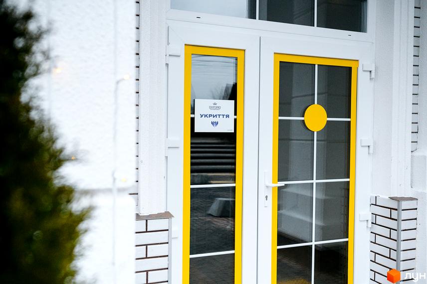 Вхід до Житлового комплексу OXFORD з білими стінами та жовто-білими скляними дверима, на яких видно табличку 