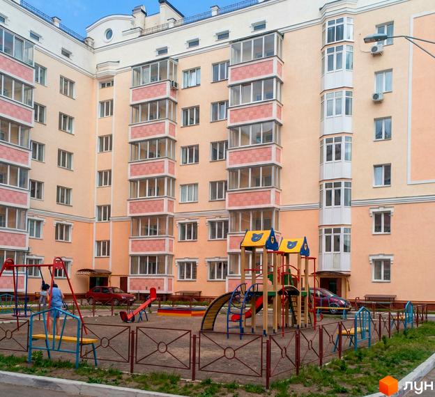 Багатоповерховий житловий комплекс персикового кольору з білими балконами та численними вікнами. У дворі облаштований дитячий майданчик.