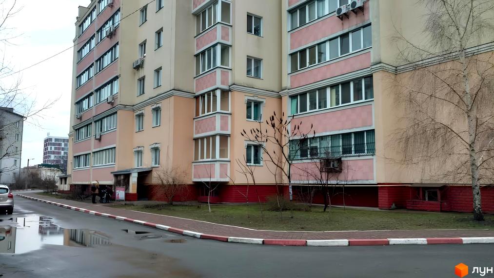 Багатоповерховий житловий будинок з бежево-рожевим фасадом та численними вікнами. Прибудинкова територія має доріжку, зелений газон та дерева.