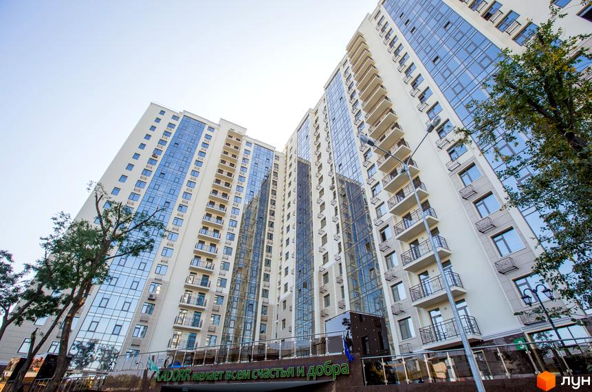 Висотна будівля ЖК Шоста Перлина з великою кількістю вікон та балконів. На передньому плані видно зелень та вивіску з написом.