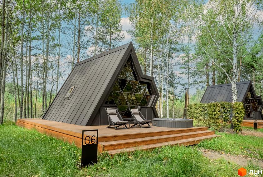 Будинок у формі трикутника з великим вікном та терасою, оточений деревами. Поруч розташована гідромасажна ванна та сусідні будинки.