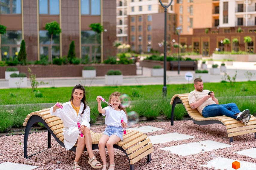 Зовнішній вигляд житлового комплексу KADORR City з зеленими насадженнями та зоною для відпочинку. Мати з дочкою грають на лавках, чоловік користується телефоном.