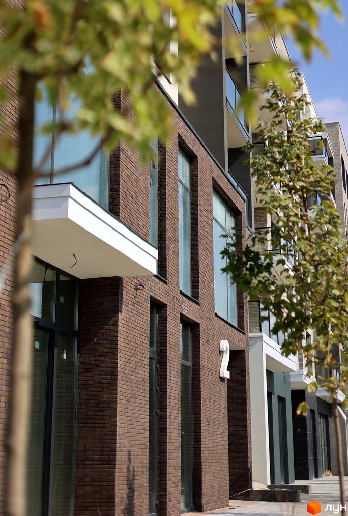 Зовнішній вигляд будинку ЖК SKY CITY з цегляною фасадною обробкою та великими вікнами. Прибудинкова територія озеленена деревами.