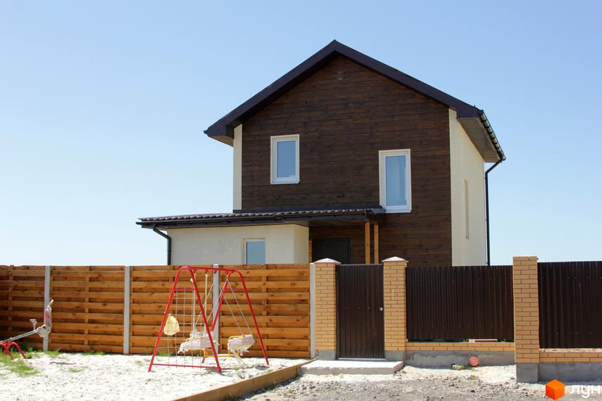 Будинок у Котеджному містечку Приозерний має темно-коричневий дерев'яний фасад та світлі стіни. На подвір'ї розташовано дитячий майданчик з гойдалками та огорожа.
